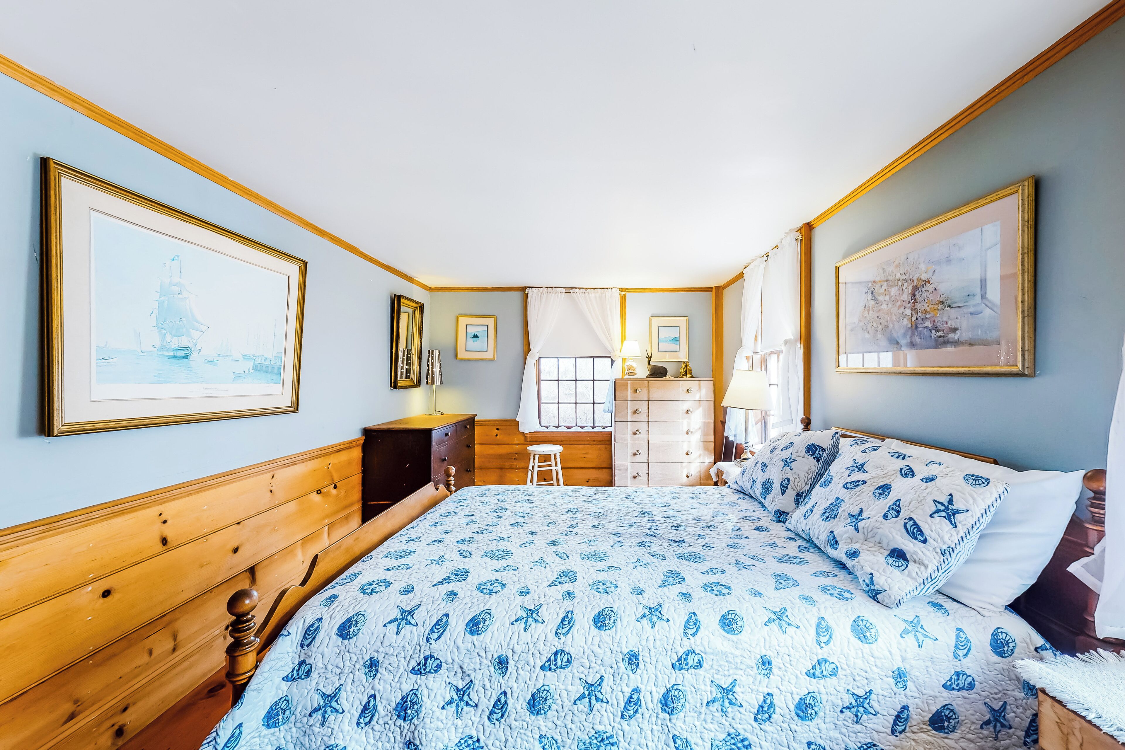 A bedroom with a wooden headboard, a bed with a blue and white patterned bedspread, a dresser, a nightstand, and two framed pictures on the wall.
