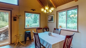 A dining area with a table set for four, wooden chairs, a chandelier, and large windows with a view of trees. - Deck & Mtn Views! Family A-frame Cabin Near Stream (Bailey)