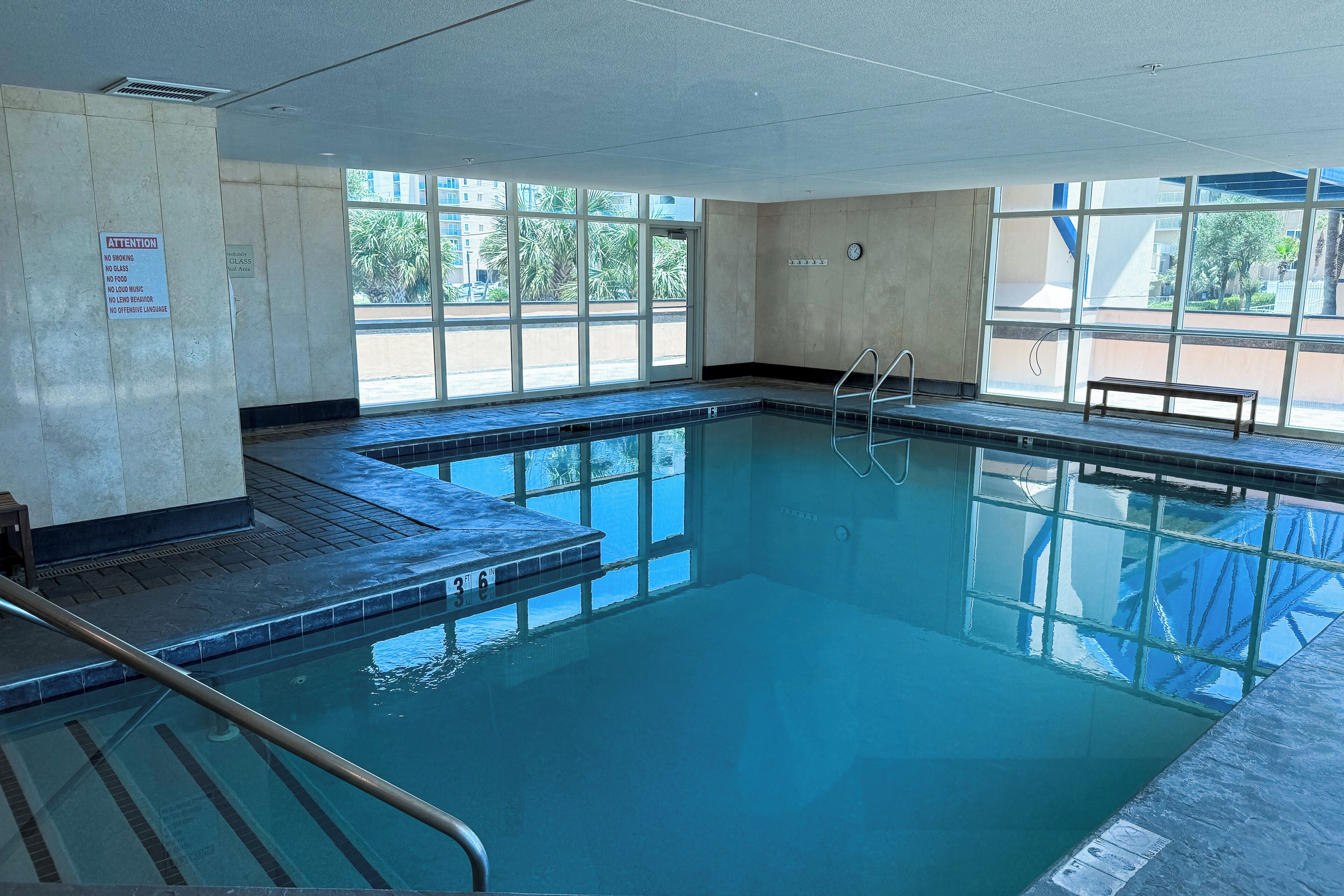 Une piscine intérieure avec des parois en verre transparent, un banc et un panneau indiquant qu’il est interdit de fumer, de consommer des boissons alcoolisées ou d’écouter de la musique à volume élevé.
