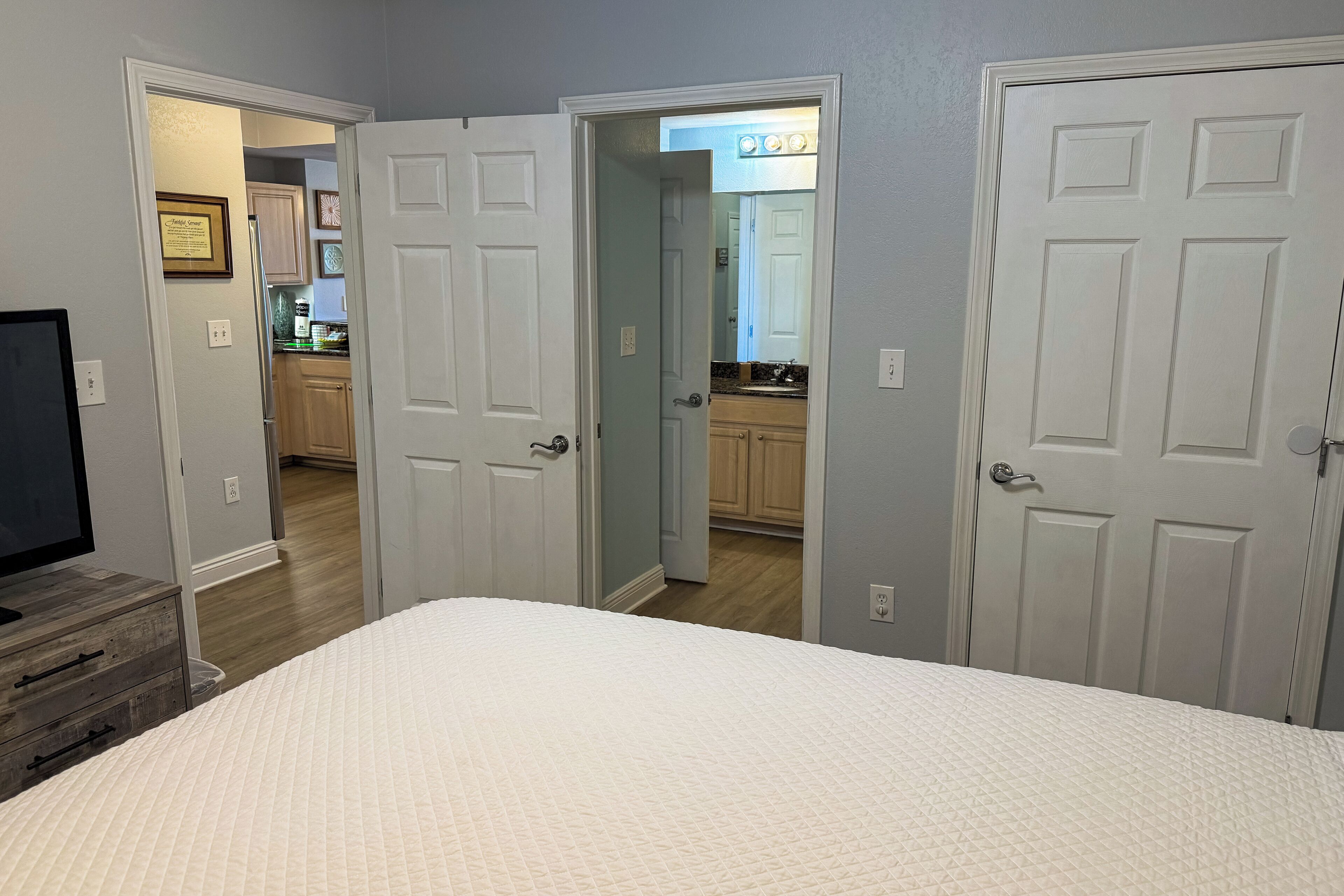 A bedroom with a large bed, a dresser, and a TV. There are three doors leading to other rooms, including a kitchen.