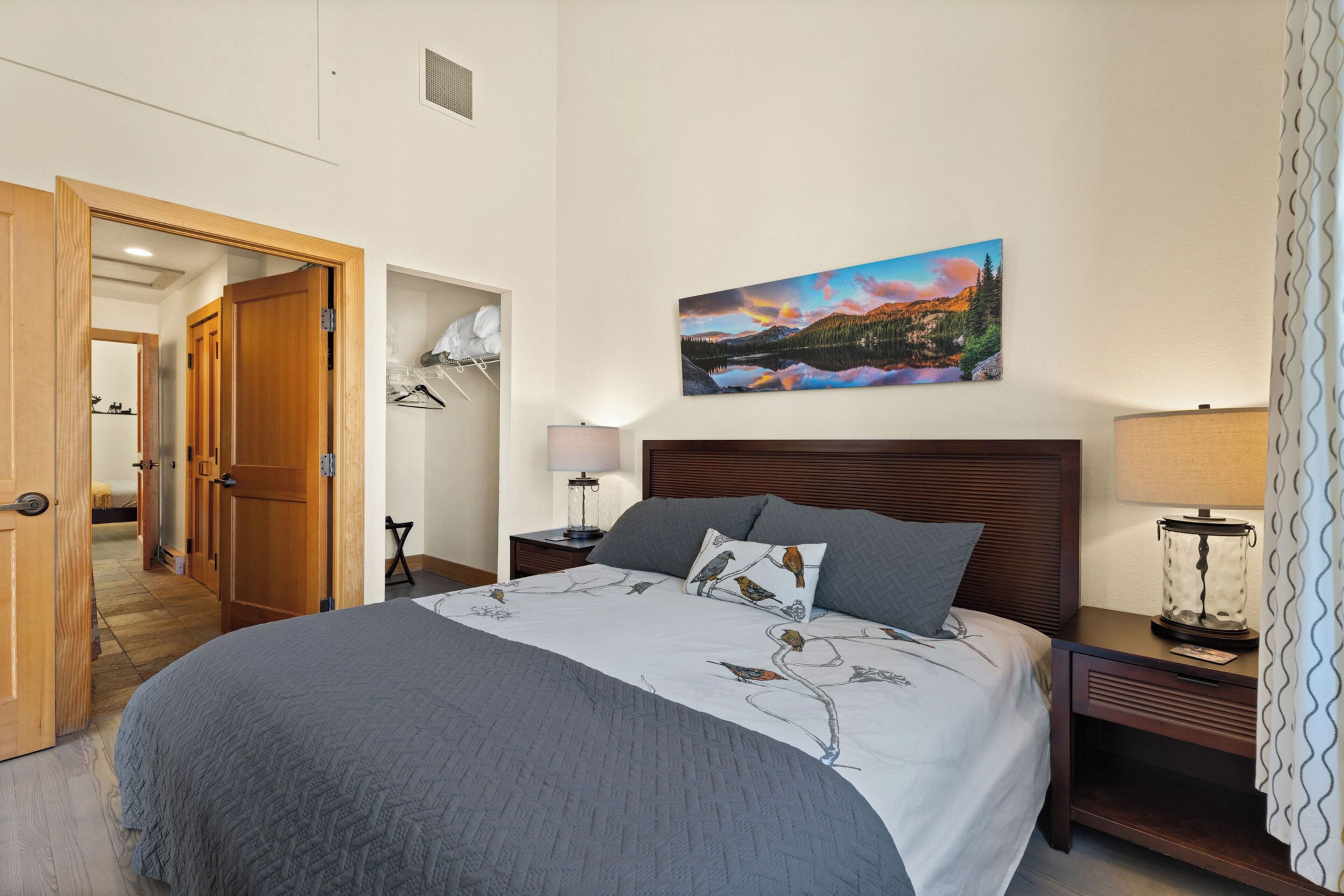 A hotel room with a large bed, a nightstand with a lamp, a wall-mounted painting, and a door leading to another room.