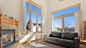 Un salon chaleureux avec une cheminée, un canapé en cuir et de grandes fenêtres donnant sur une maison et des montagnes.