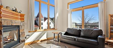Un salon chaleureux avec une cheminée, un canapé en cuir et de grandes fenêtres donnant sur une maison et des montagnes.