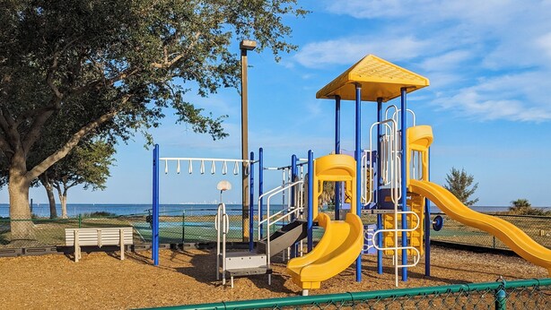 Ein Spielplatz mit einer gelben Rutsche und blauen Spielgeräten, umgeben von einem Zaun und Bäumen.
