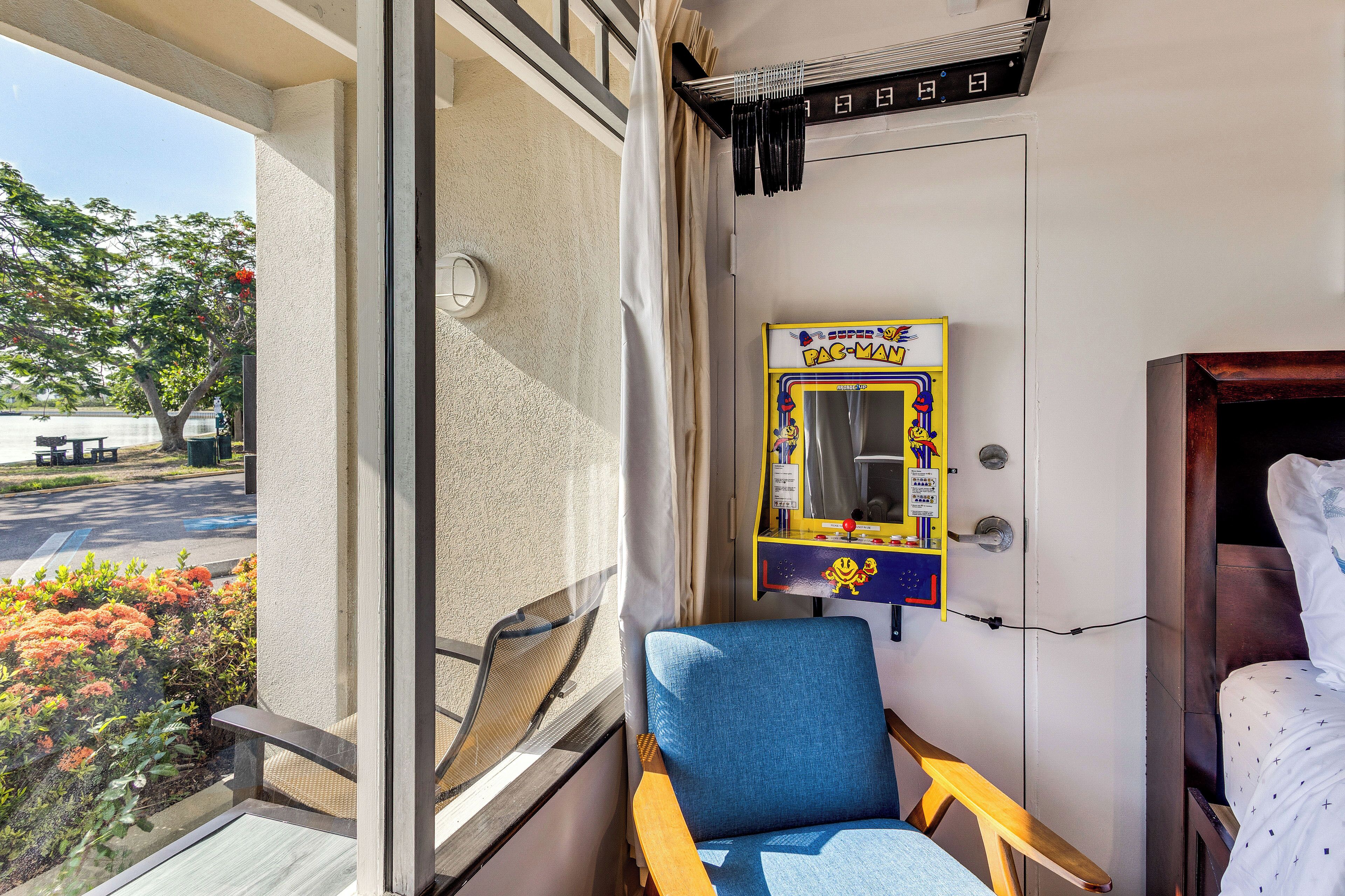 A room with a window, a chair, a bed, a wall-mounted game machine, and a door.
