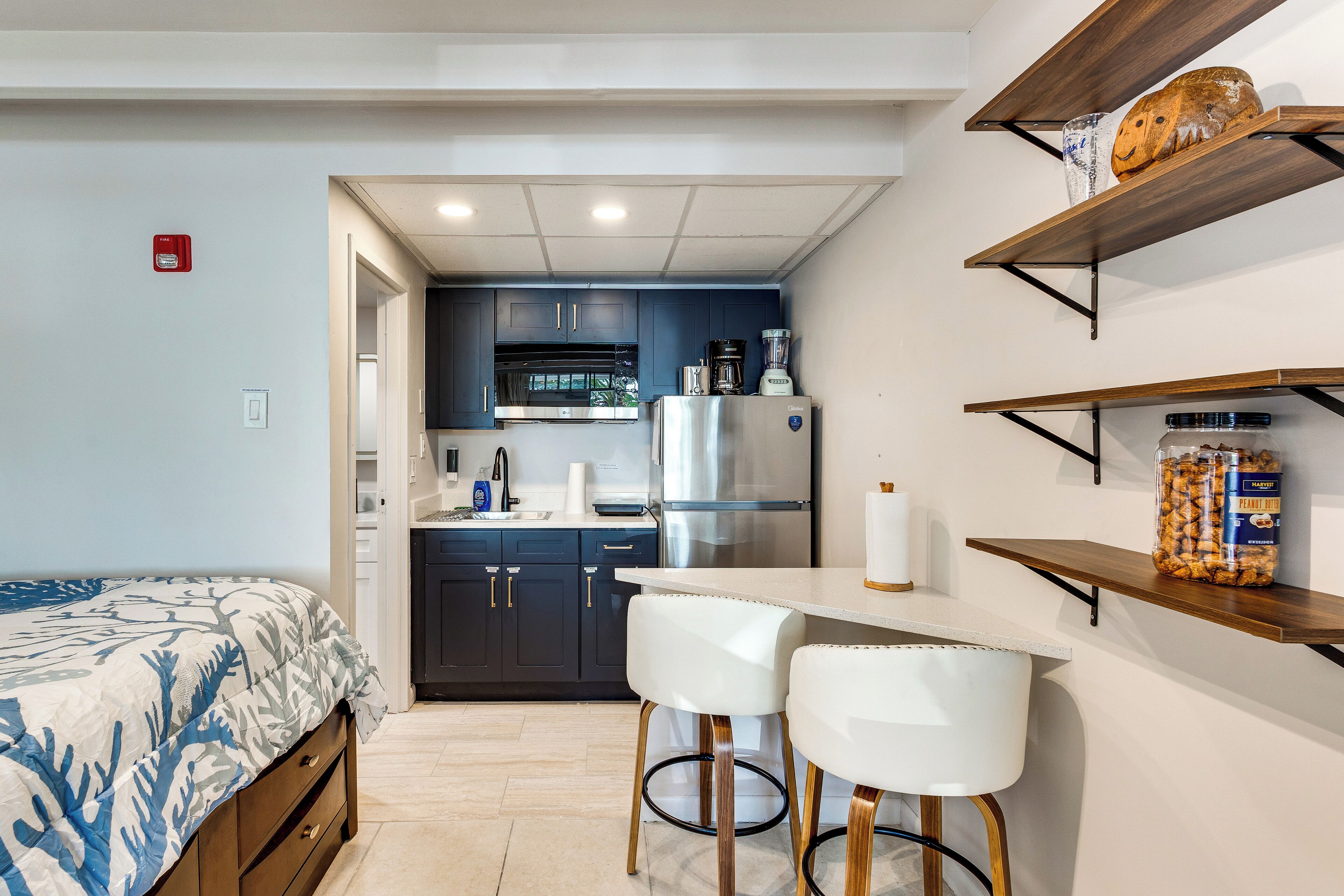 A compact kitchen with dark cabinets, a refrigerator, and a coffee maker. There are two white bar stools and a small table.