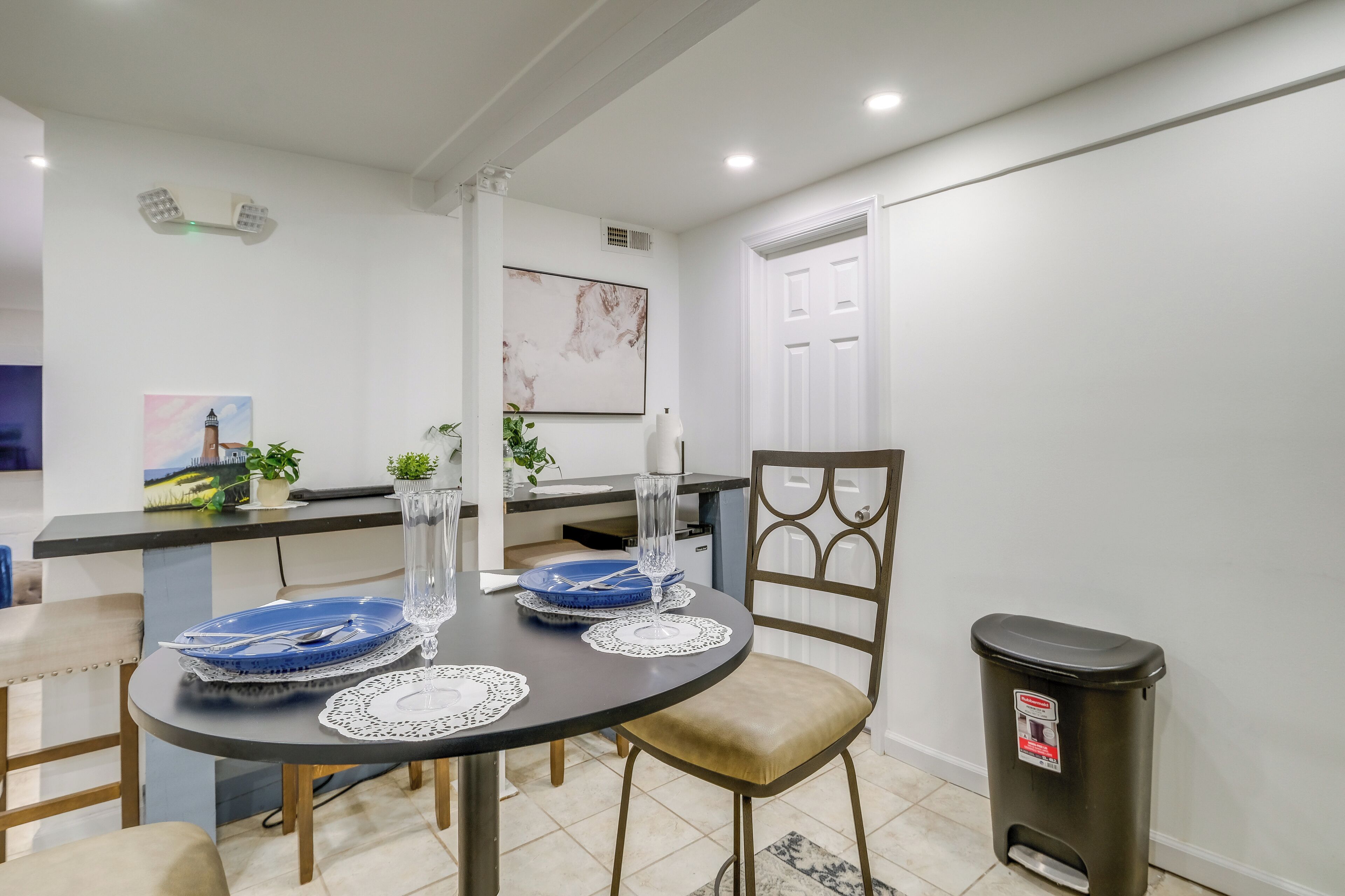 A modern kitchen with a dining table set for four, a trash bin, and a white door in the background.