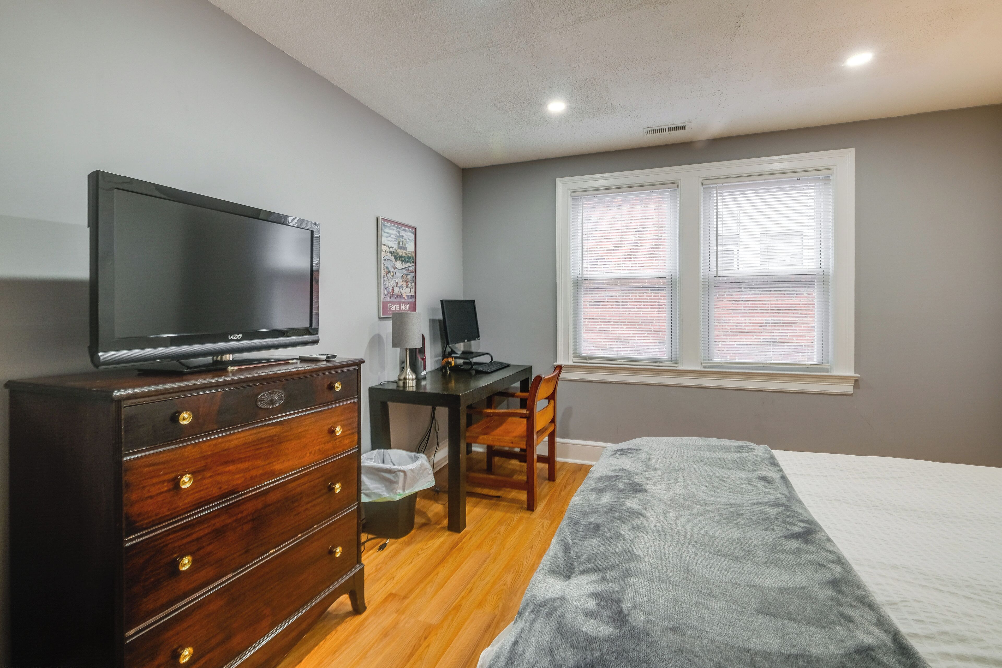 Un dormitorio con cama, cómoda, televisión, escritorio, silla y ventana con persianas.