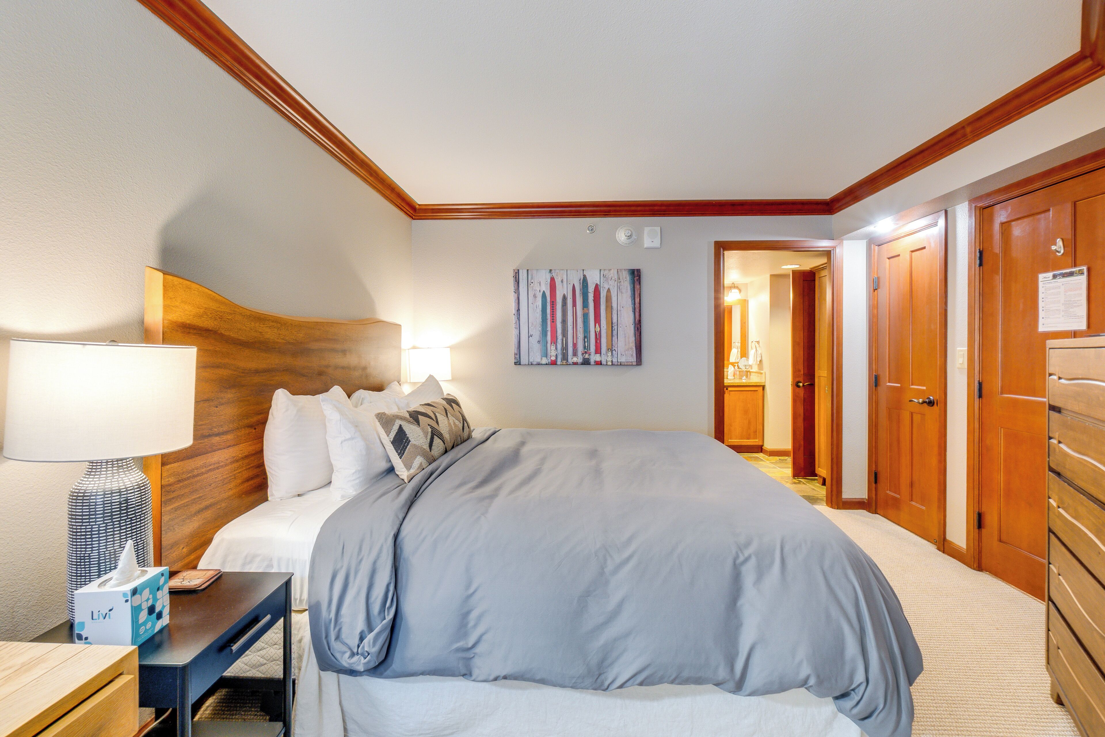 Ein Hotelzimmer mit einem großen Bett, einem Holzkopfteil, einem Nachttisch mit Lampe, einem an der Wand hängenden Kunstwerk und einem Holzschrank.