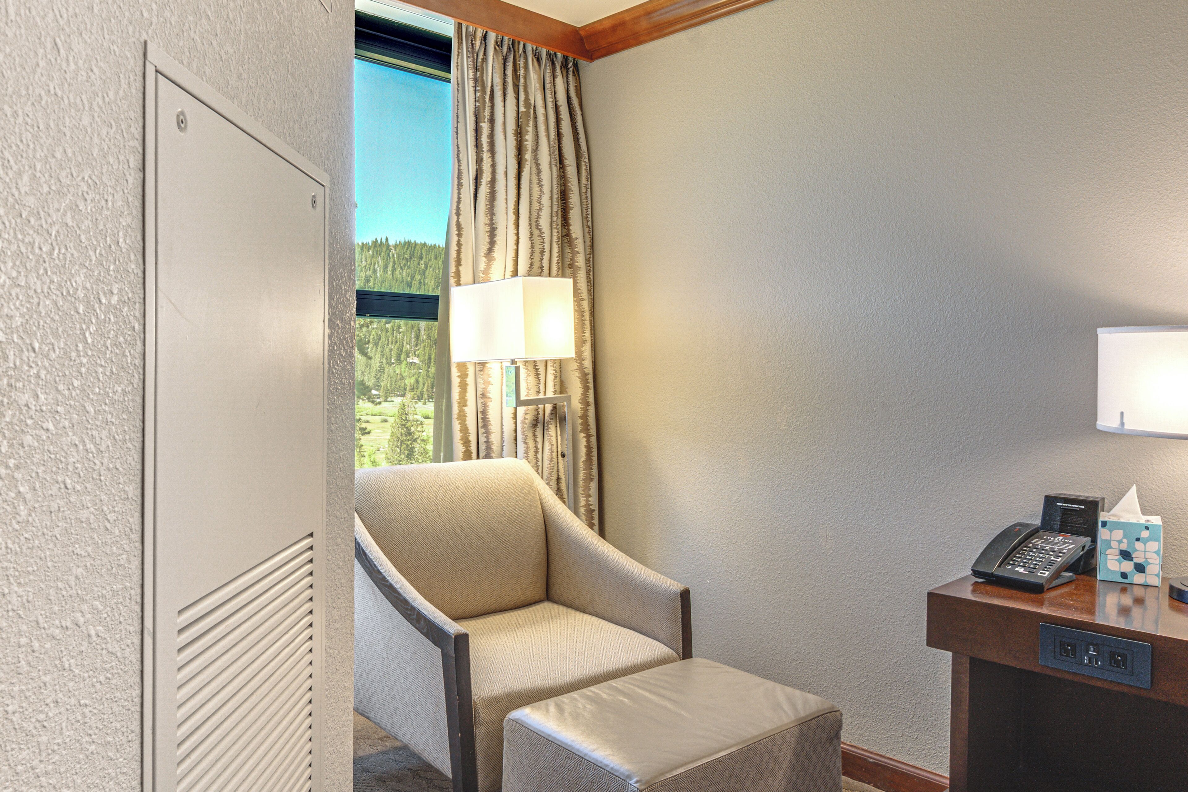 A hotel room with a chair, a desk with a phone and tissues, and a window with curtains.