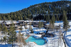 A resort with a swimming pool surrounded by snow, buildings, and a forested mountain backdrop.