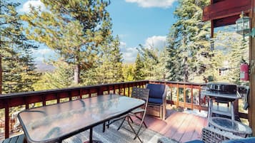 A wooden deck with a glass table and chairs, surrounded by tall trees and a mountain view.