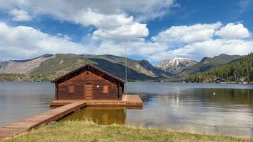 A wooden cabin on a dock by a lake with mountains in the background.