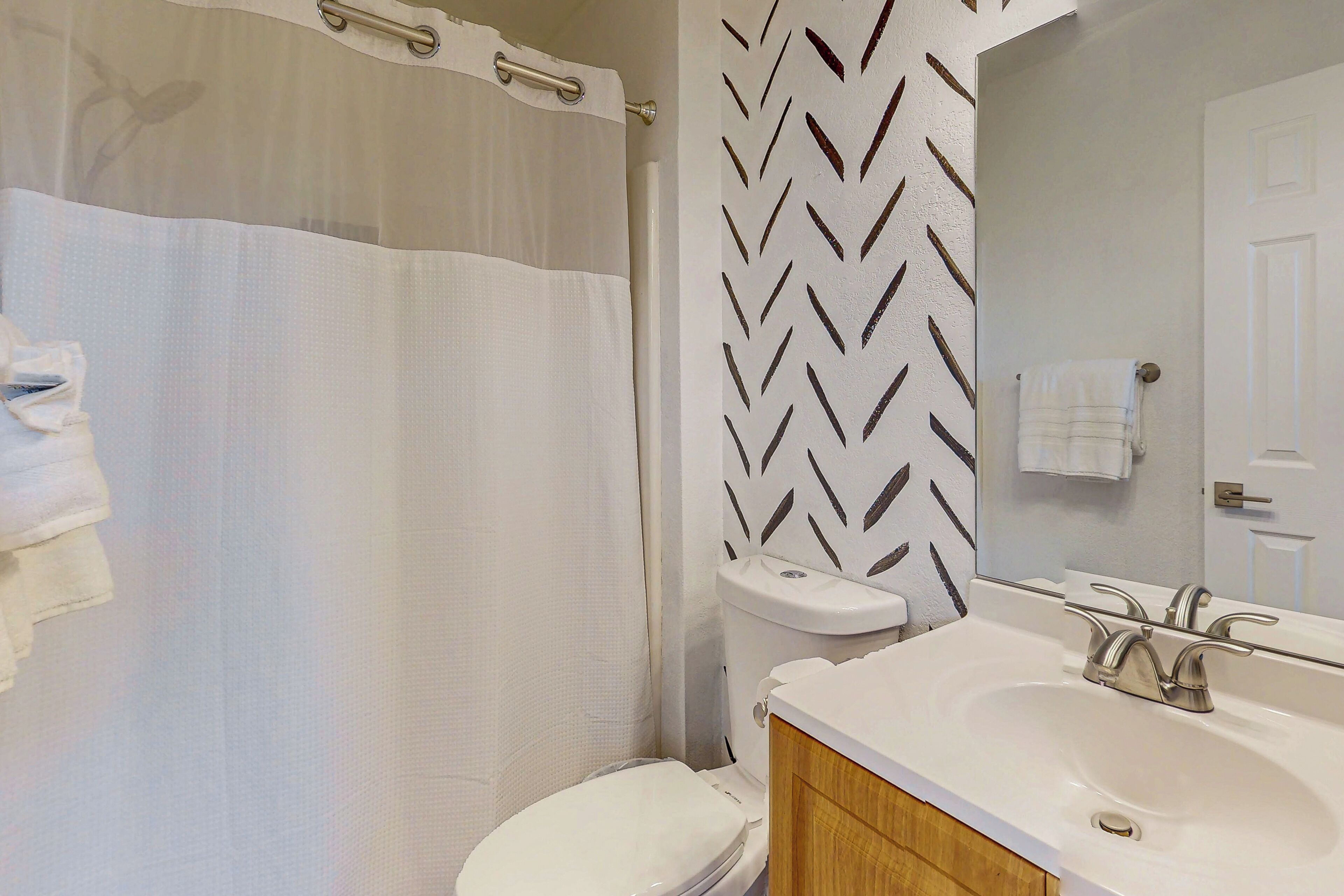 Une salle de bain avec des toilettes blanches, un rideau de douche, un lavabo avec un miroir et un mur à motifs.
