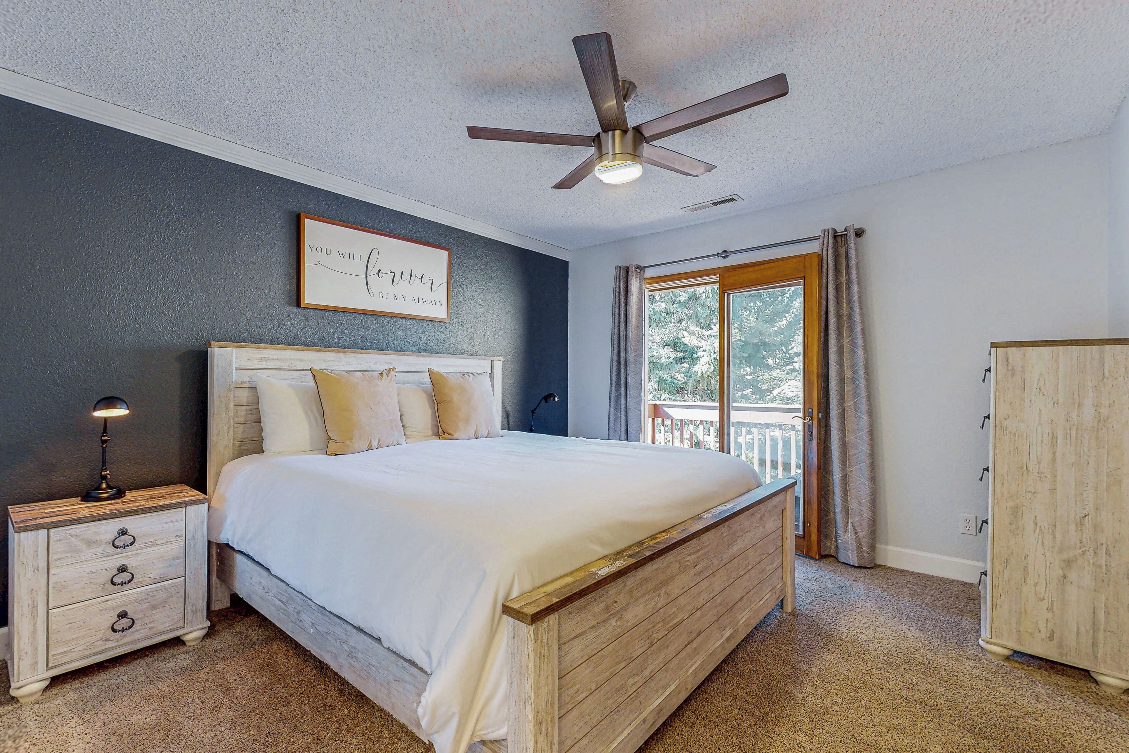 A bedroom with a bed, a nightstand, a ceiling fan, a window with curtains, and a view of the outdoors.