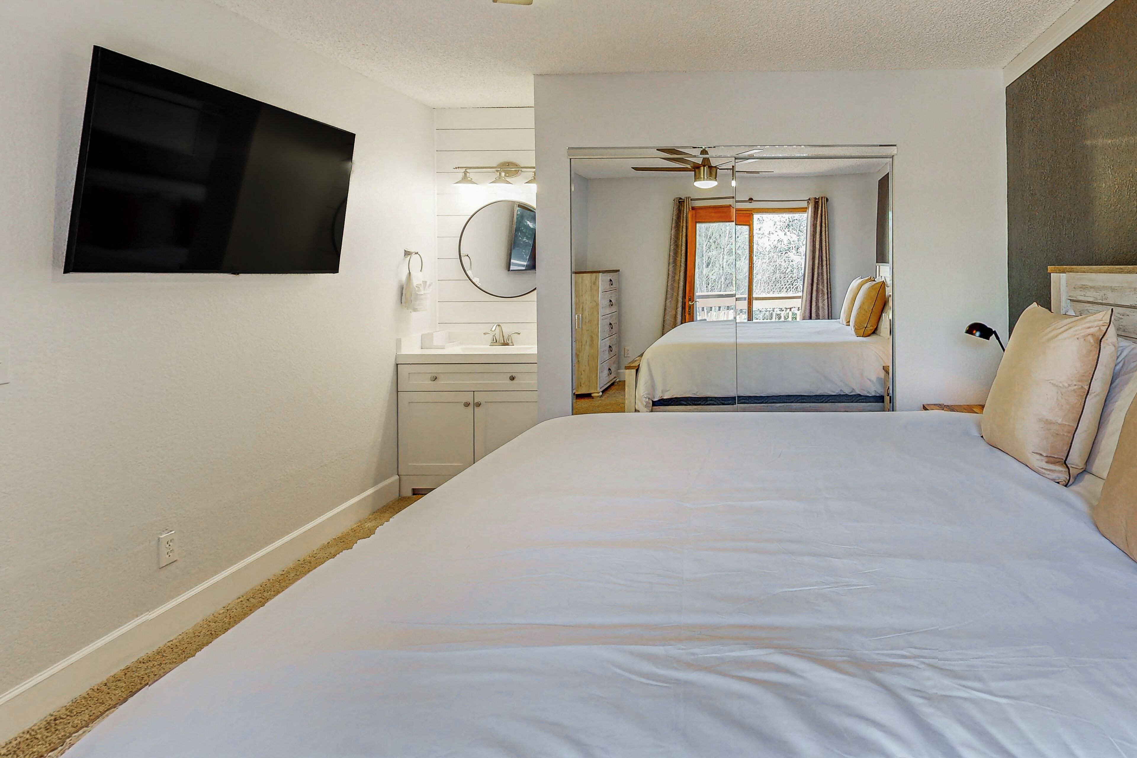 A bedroom with a large bed, a TV mounted on the wall, a ceiling fan, and a window with curtains.