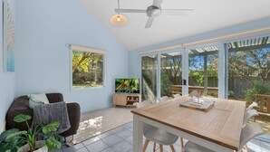 A spacious living area with a large sliding glass door leading to a garden, a ceiling fan, and a television on a wooden stand. - Aldebaran Road Beach House - perfect family coastal getaway, your search ends here (Ocean Grove)
