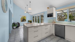 A modern kitchen with a large island, white cabinetry, and a built-in microwave. There is a window with a view of trees and a sign on the window sill. - Aldebaran Road Beach House - perfect family coastal getaway, your search ends here (Ocean Grove)