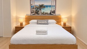 A neatly made bed with a white comforter, a pillow with the word 'beach,' and a view of a beach scene through a window. - Aldebaran Road Beach House - perfect family coastal getaway, your search ends here (Ocean Grove)