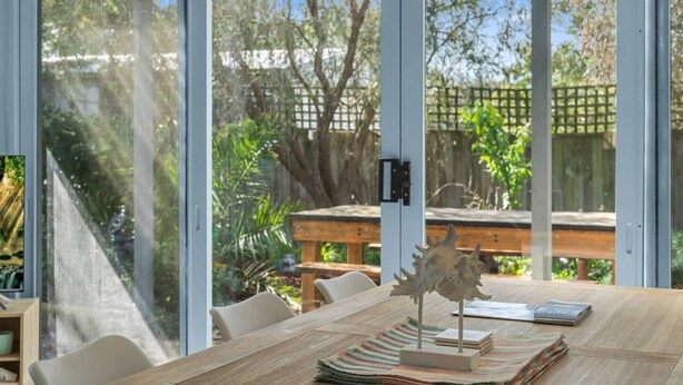 A dining area with a wooden table, chairs, and a view of a garden through large windows. - Aldebaran Road Beach House - perfect family coastal getaway, your search ends here (Ocean Grove)
