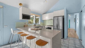A modern kitchen with a central island, bar stools, a stainless steel refrigerator, and a window providing natural light. - Aldebaran Road Beach House - perfect family coastal getaway, your search ends here (Ocean Grove)