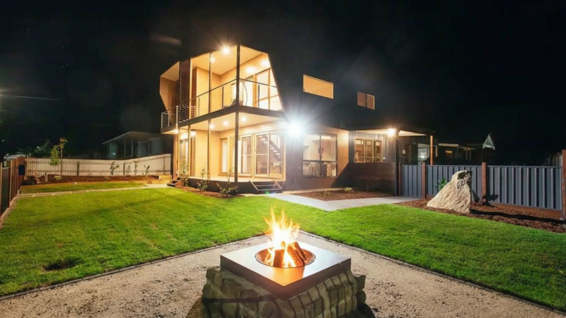 A modern two-story house with a well-lit interior and a fire pit in the yard.