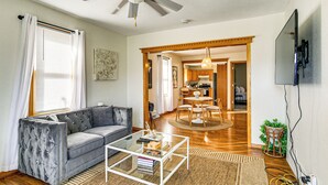 A living room with a grey sofa, a glass coffee table, a ceiling fan, and a kitchen visible through an open doorway. - Pet-friendly Home 2 Mi to Downtown Des Moines! (Des Moines)