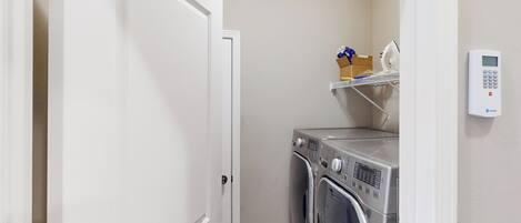 A laundry room with a washing machine and dryer, a shelf with folded clothes, and a digital thermostat on the wall.