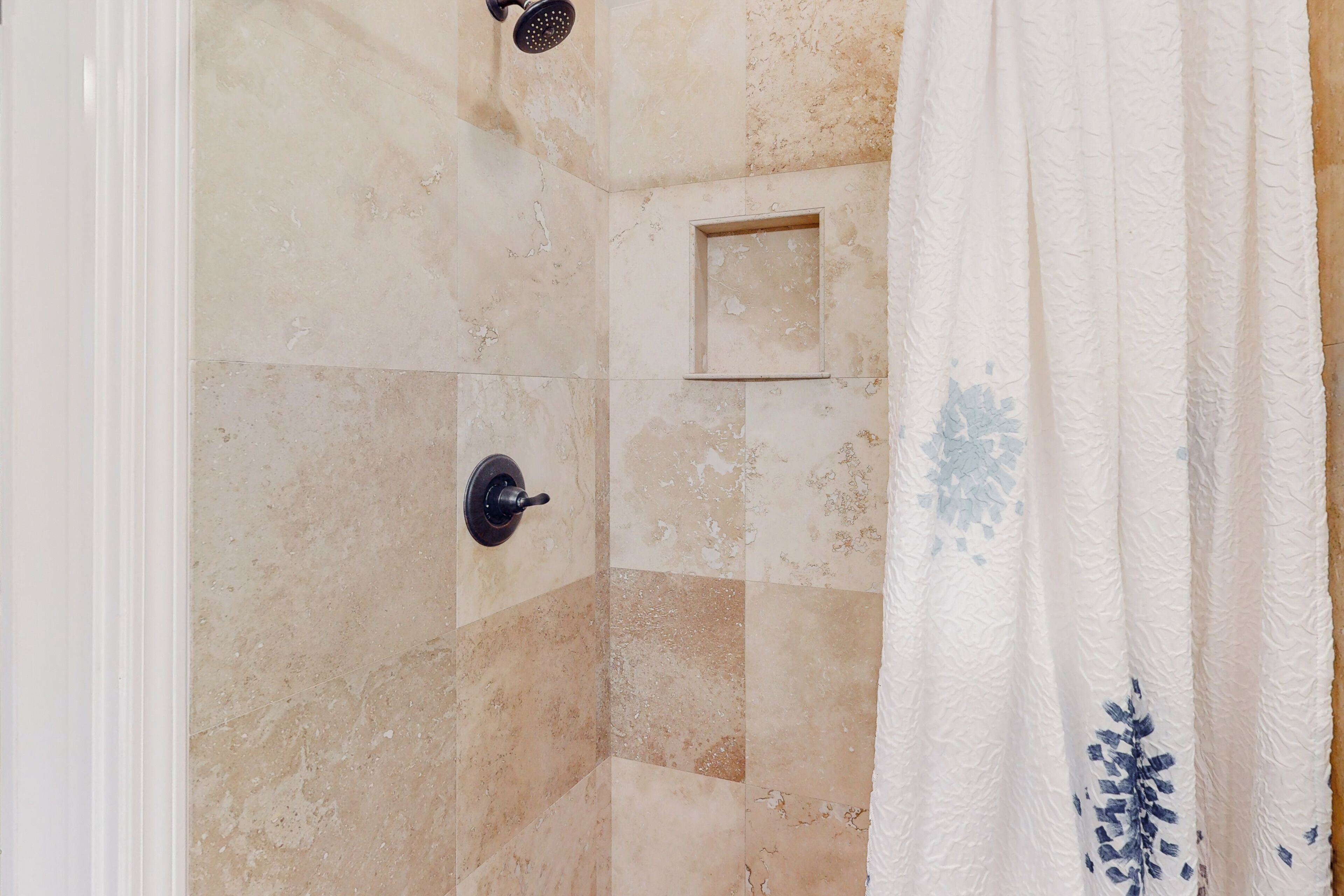 A shower area with beige tiles and a white shower curtain with blue floral patterns.