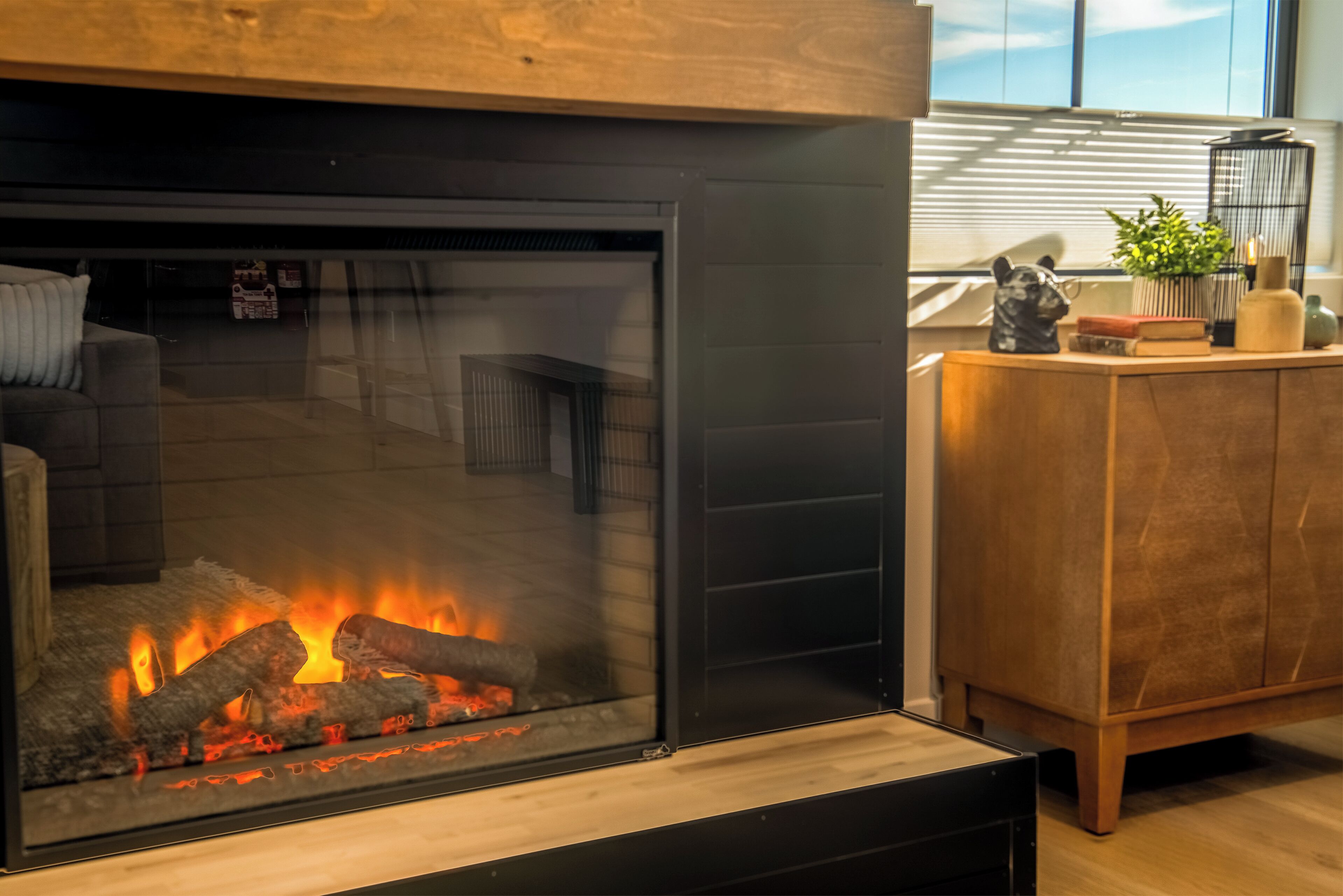 A modern fireplace with visible logs burning, a wooden cabinet to the right, and a window with blinds in the background.