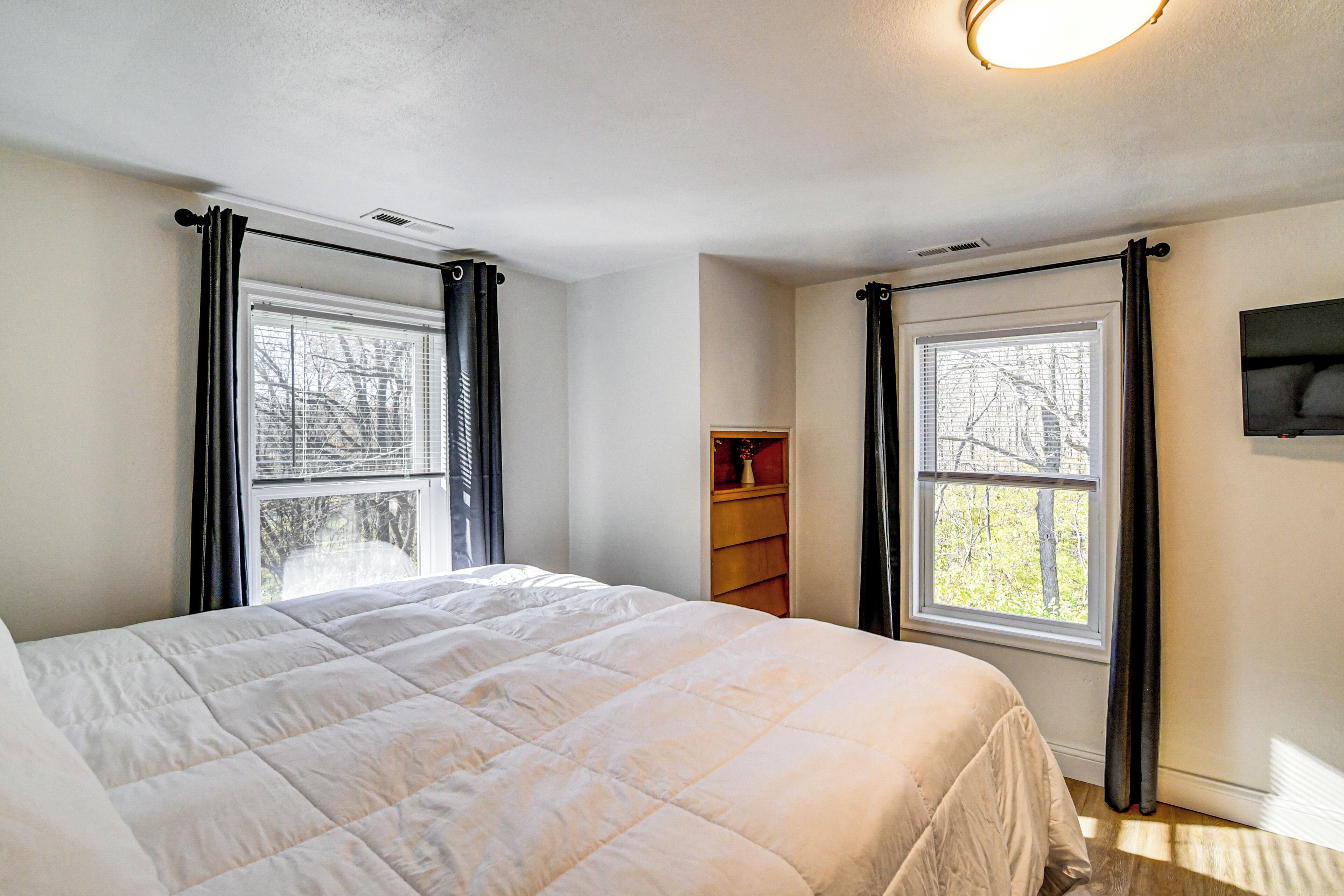 A bedroom with a large bed, two windows with curtains, a nightstand, and a TV mounted on the wall.