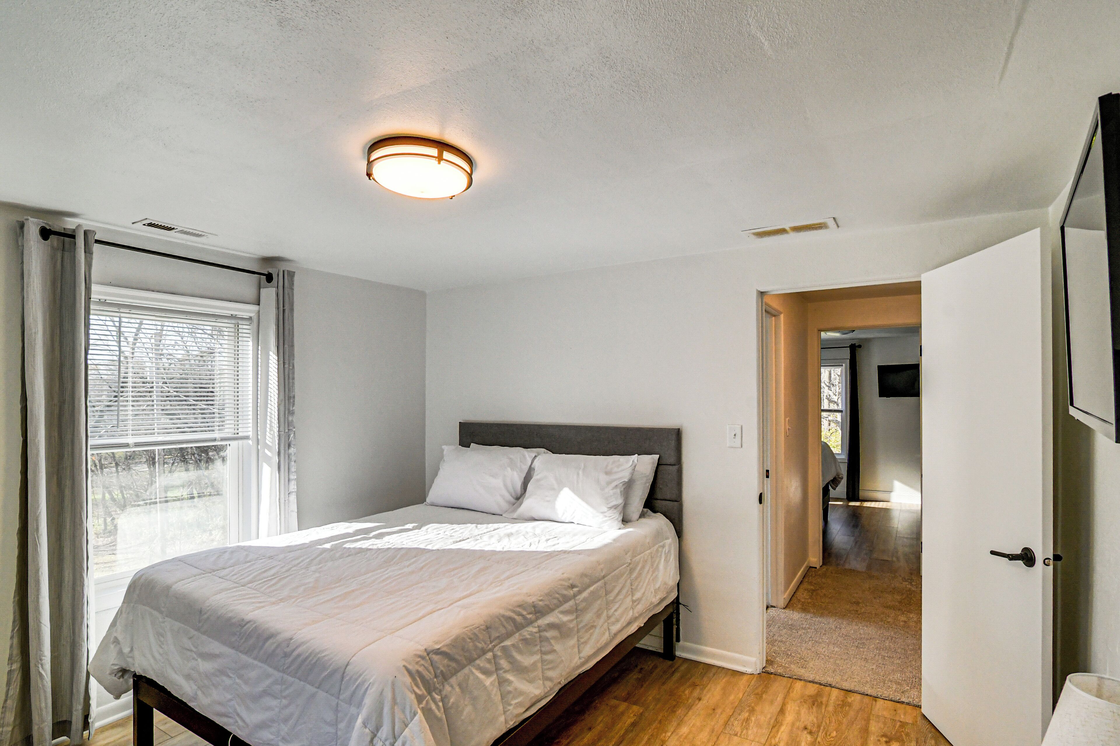 A bedroom with a bed, a ceiling light, a window with curtains, and a door leading to another room.