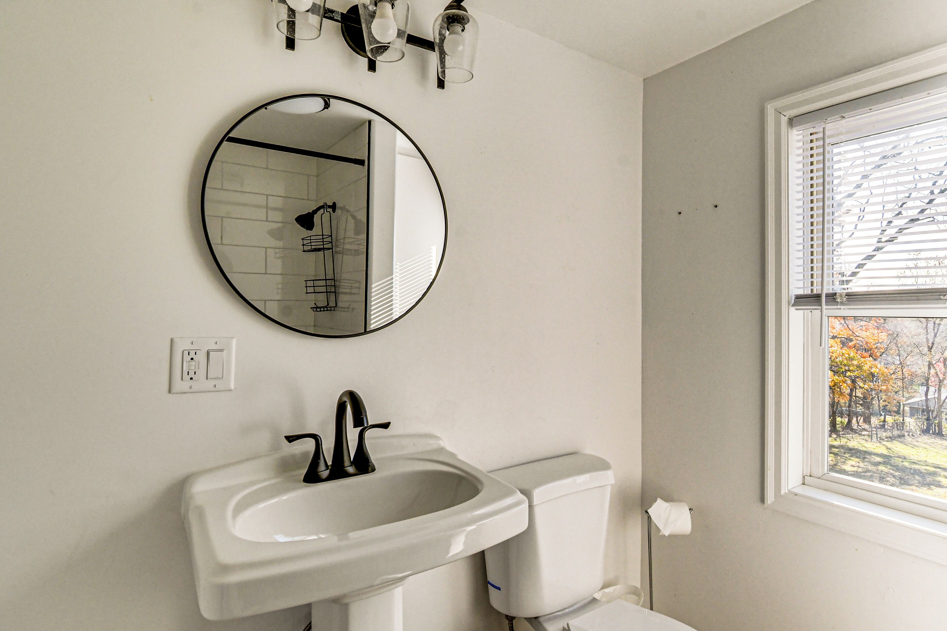 Een badkamer met een ronde spiegel, een wastafel, een toilet en een raam met zonwering.