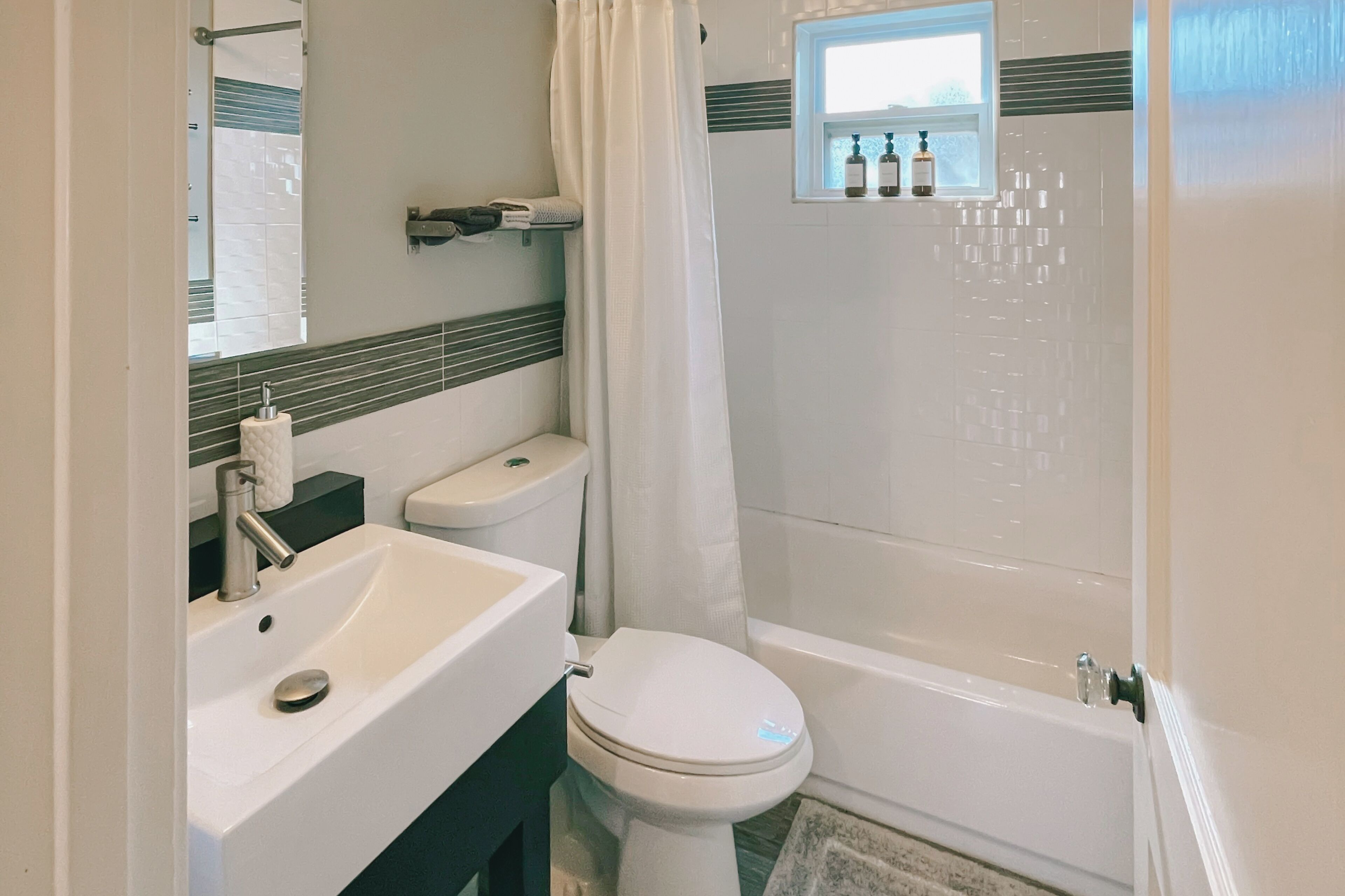 Un baño con lavamanos, inodoro y bañera blancos. Hay una ventana sobre la bañera, con botellas en el alféizar.
