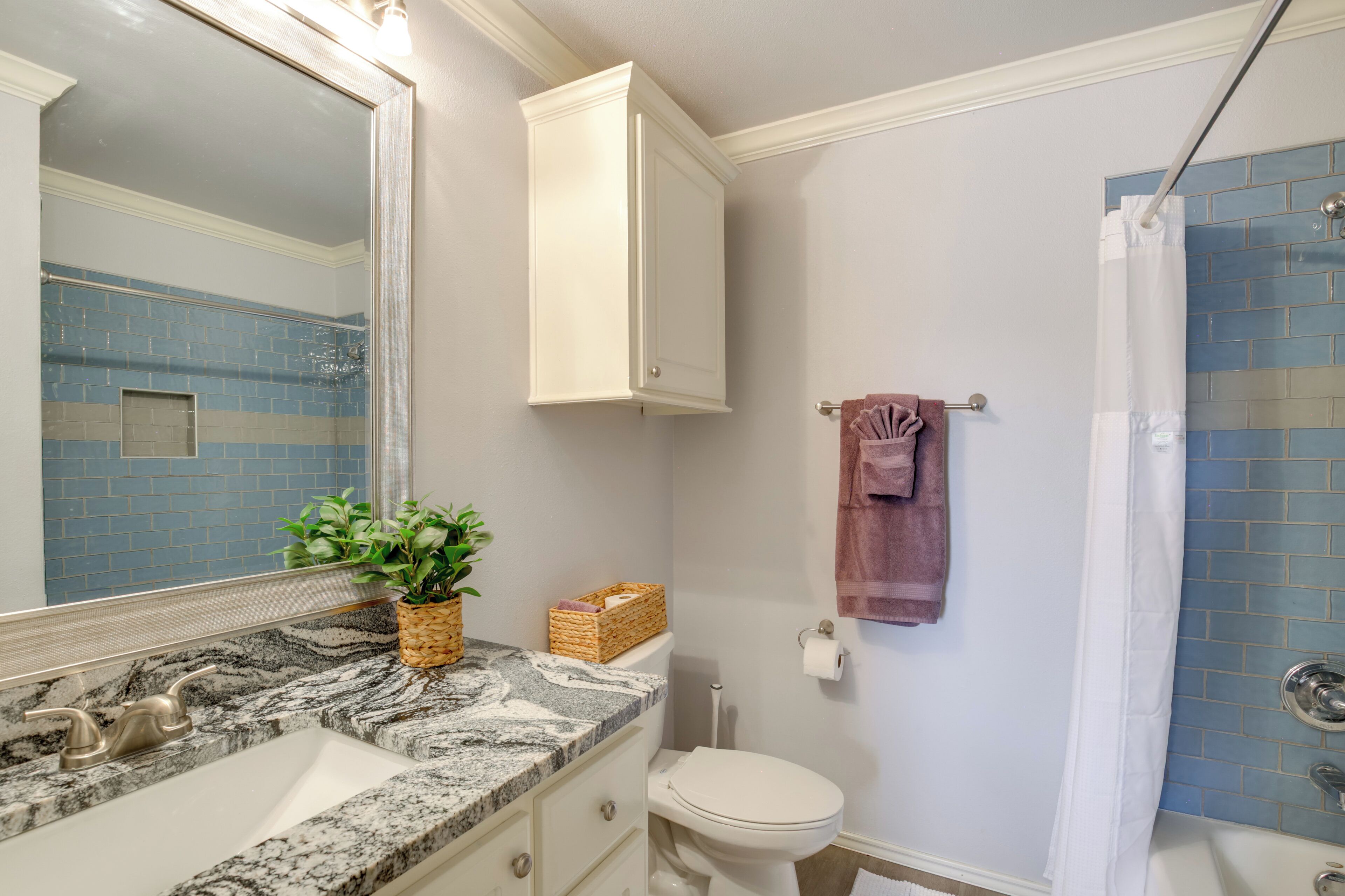 A bathroom with a marble sink, a white toilet, a shower with a blue-tiled wall, and a mirror with a cabinet above it.