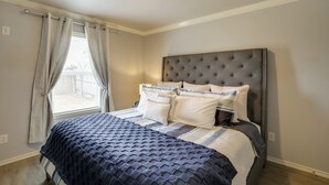 Une chambre à coucher avec un grand lit, une tête de lit capitonnée et une fenêtre avec des rideaux.