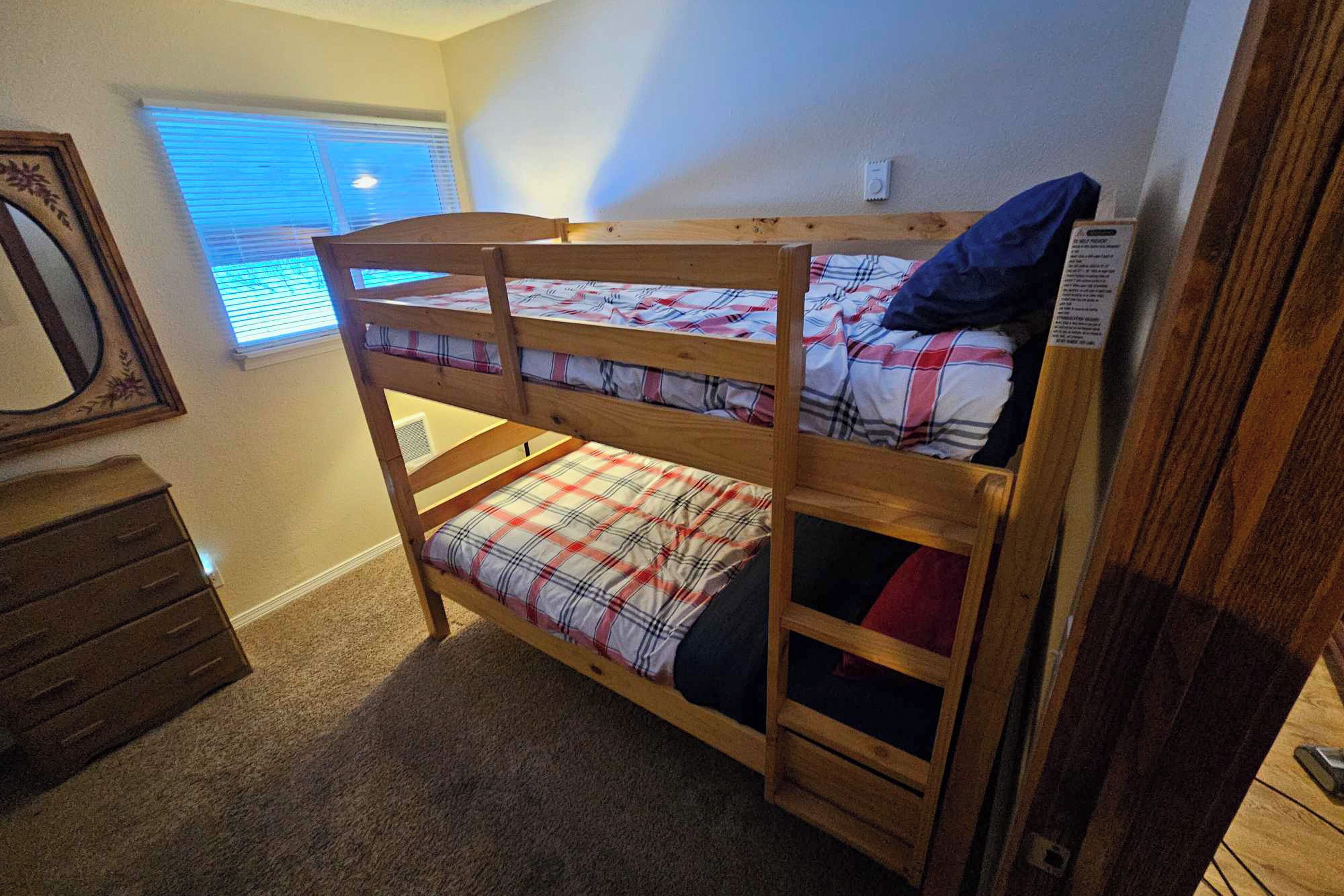 Een stapelbed met een ruitjes sprei, een houten dressoir en een raam met zonwering.
