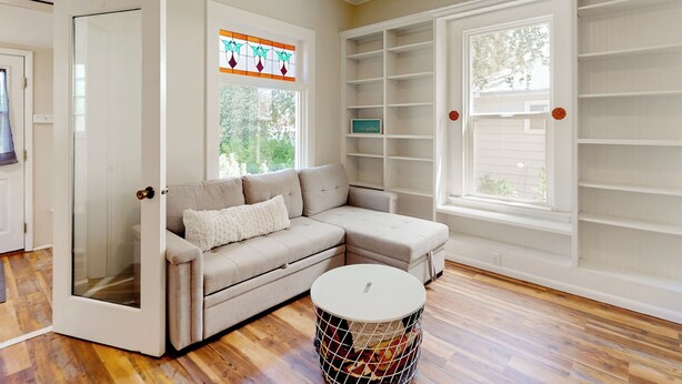 A living room with a beige sectional sofa, a round coffee table, and a large window with a stained glass design. - 2BR Home w/ Mountain View, W & D, Private Yard (Ashton)