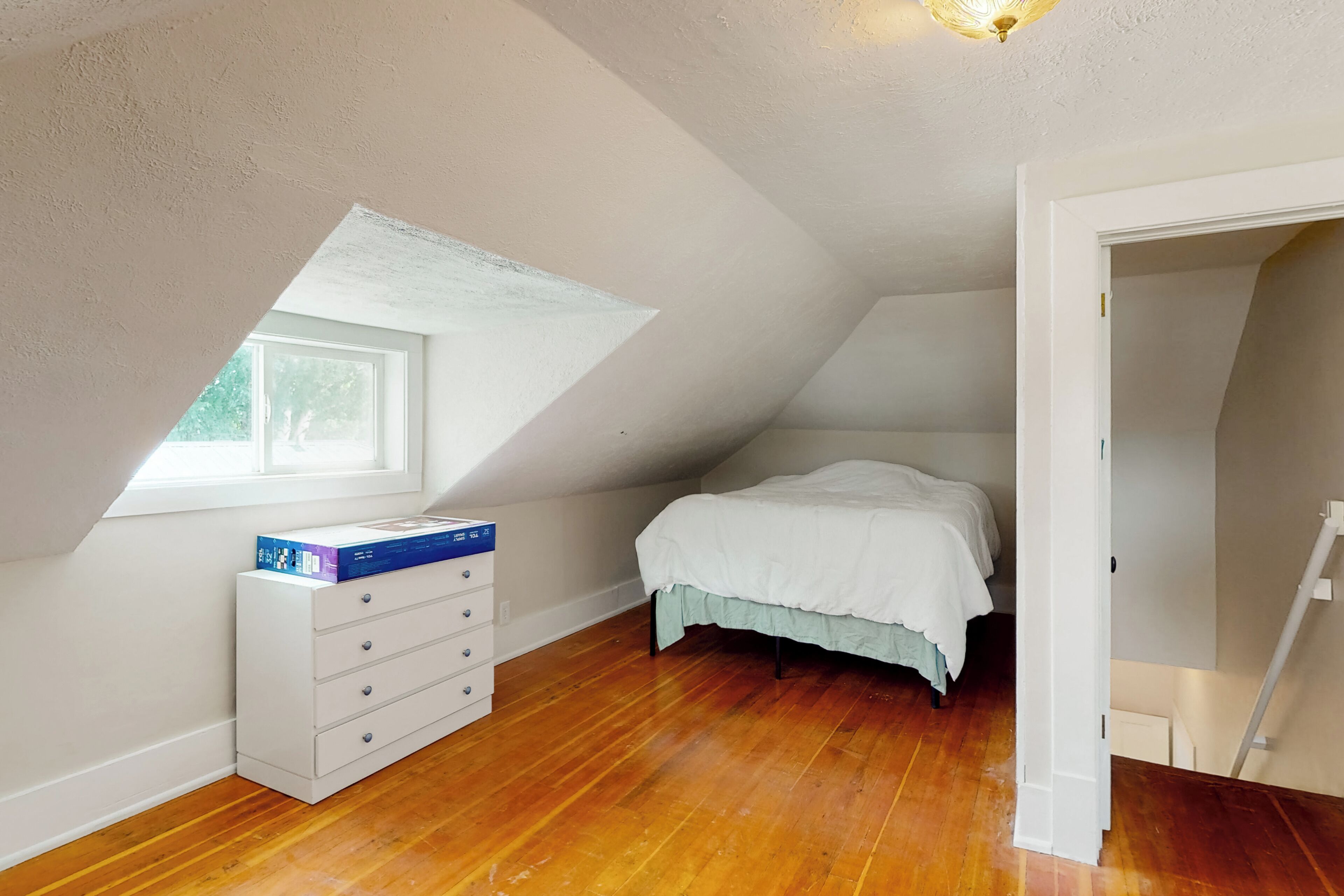 A bedroom with a sloped ceiling, a window, a bed, a dresser, and a door.