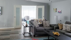 Ein modernes Wohnzimmer mit einem grauen Sofa, einem Couchtisch und einer Lampe. Eine Tür führt in einen anderen Raum, und ein Fenster ist mit Jalousien versehen.