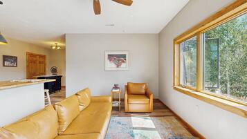 A spacious living room with a large window, a ceiling fan, a sofa, a chair, and a small table with books.