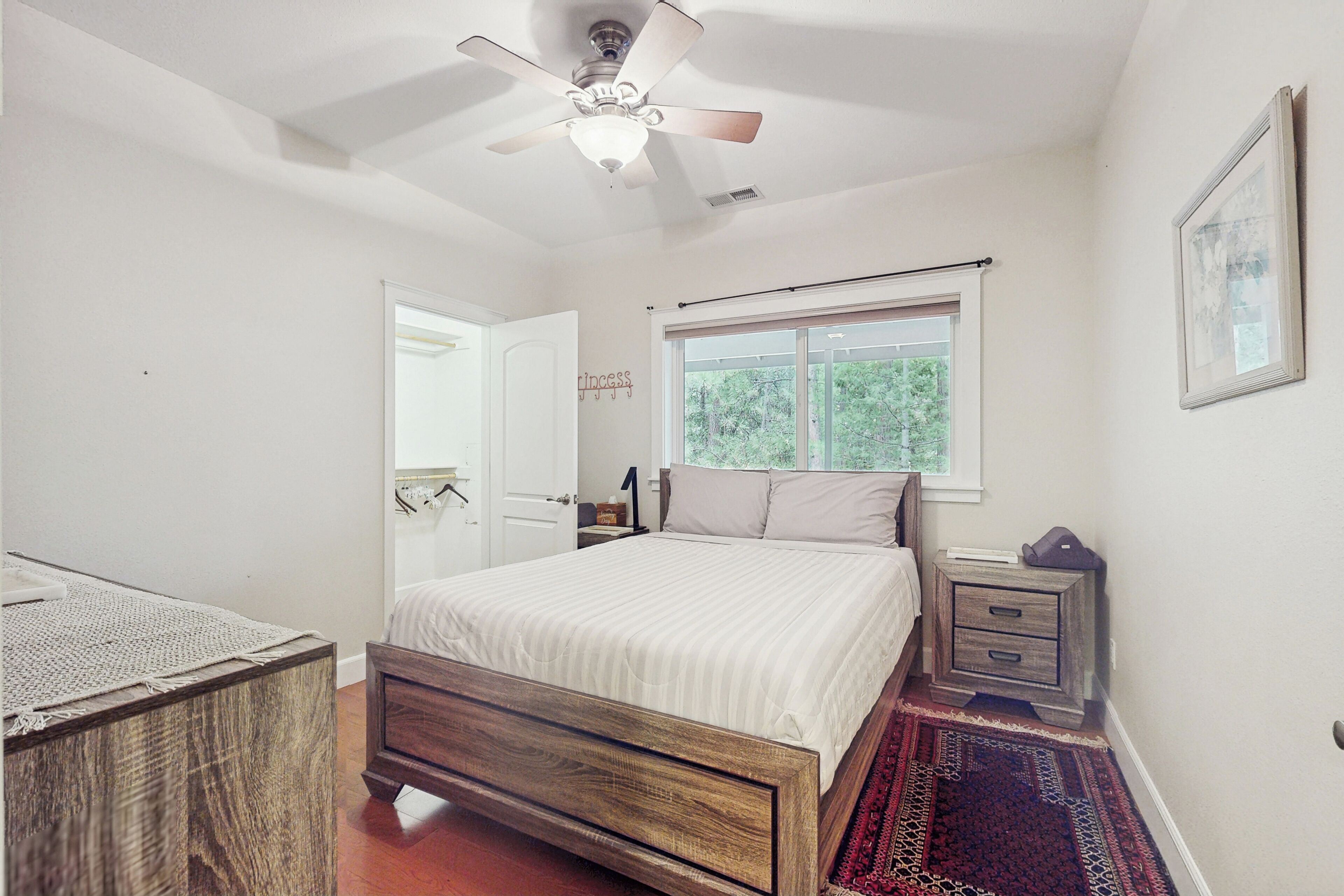 A bedroom with a wooden bed, a nightstand, a bedside table, a ceiling fan, a window with a view of trees, and a door leading to another room.