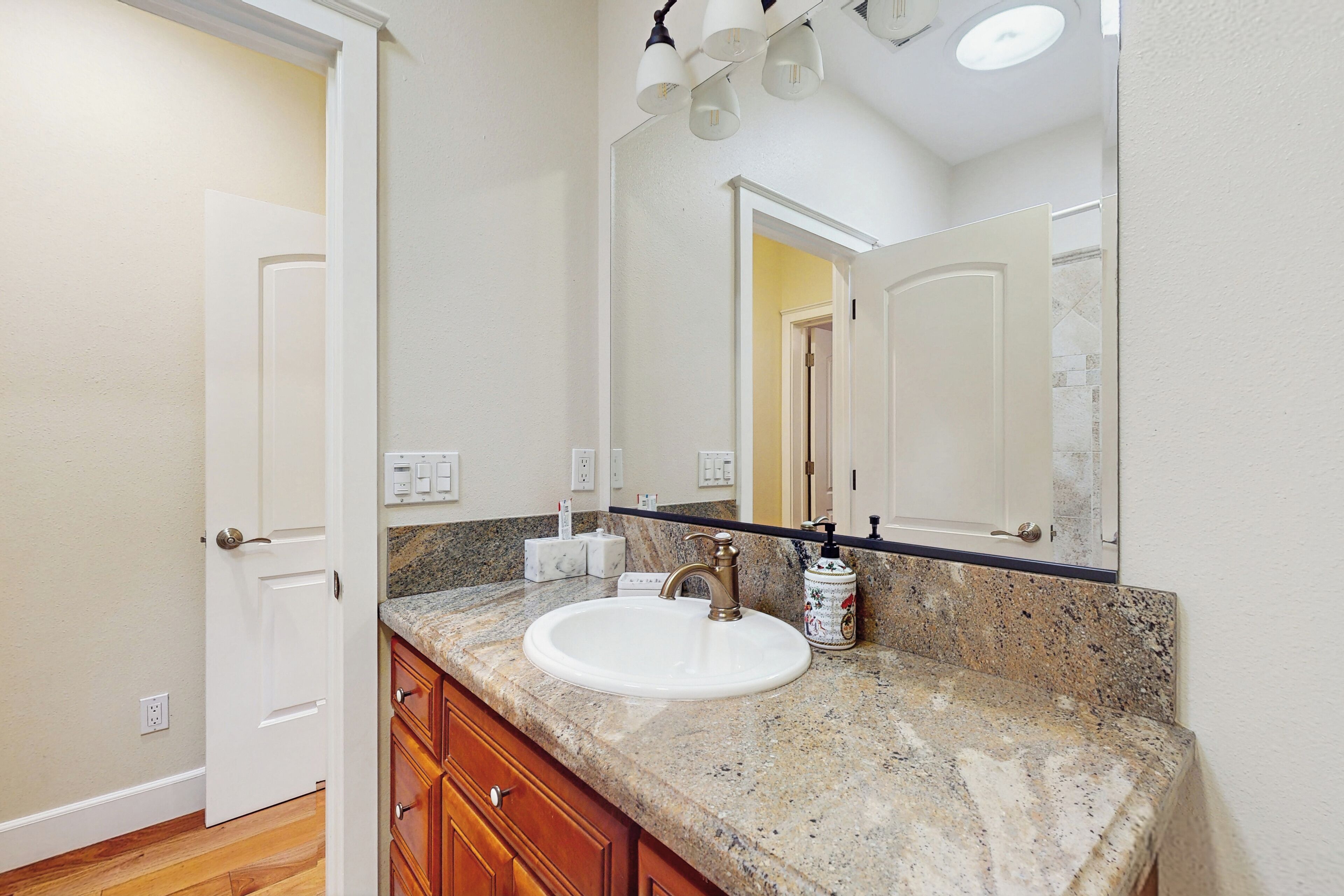 A bathroom with a white sink, granite countertop, wooden cabinets, a large mirror, and a ceiling light fixture.