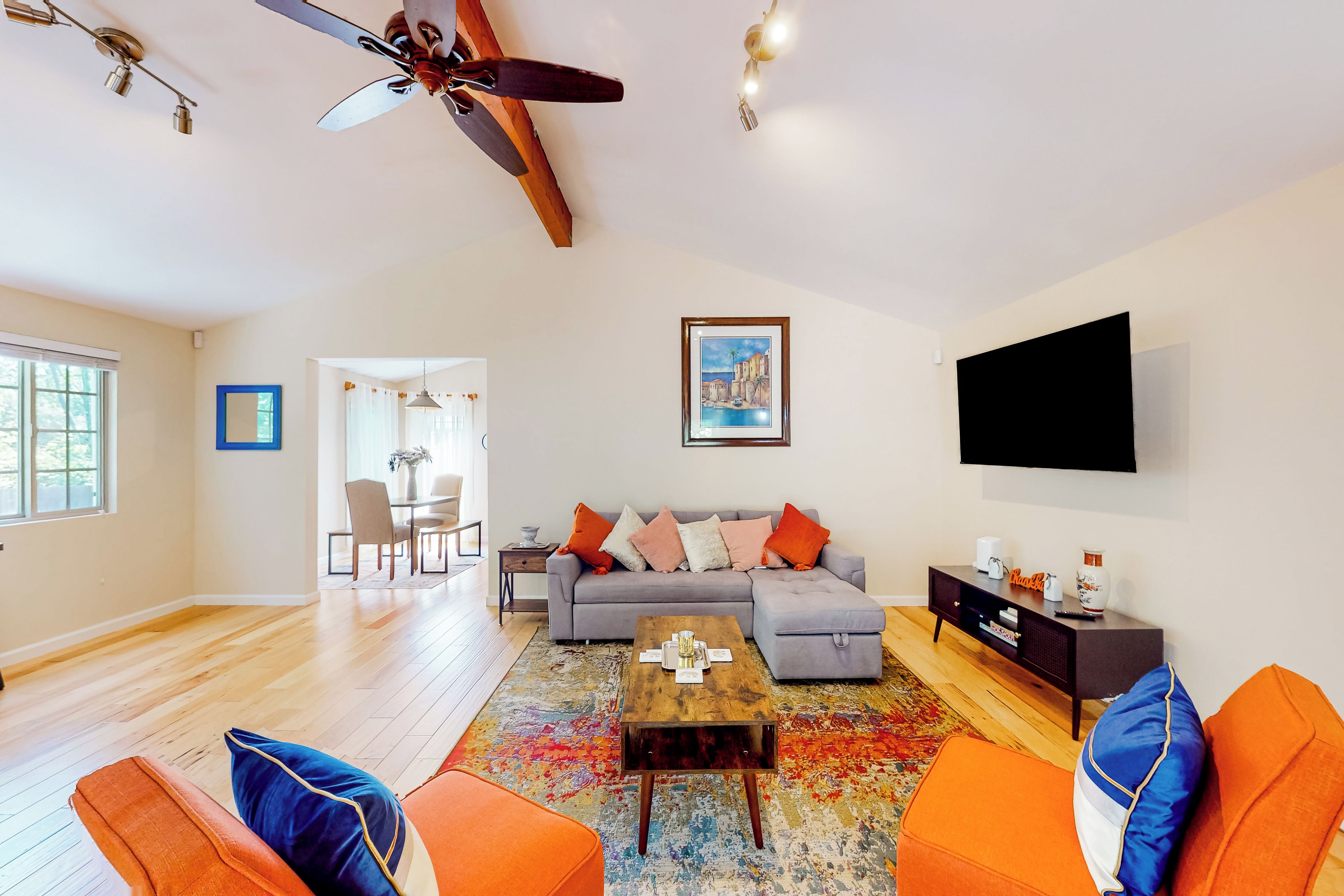 A modern living room with a sofa, coffee table, and orange chairs. There is a ceiling fan, a TV mounted on the wall, and a dining area in the background.