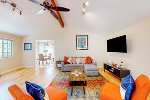 Una sala de estar moderna con un sofá, una mesa de centro y sillas color naranja. Se observa un ventilador de techo, un televisor montado en la pared y un comedor al fondo.