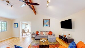 A modern living room with a sofa, coffee table, and orange chairs. There is a ceiling fan, a TV mounted on the wall, and a dining area in the background. - Centrally Located! Dog-friendly San Rafael Home (San Rafael)