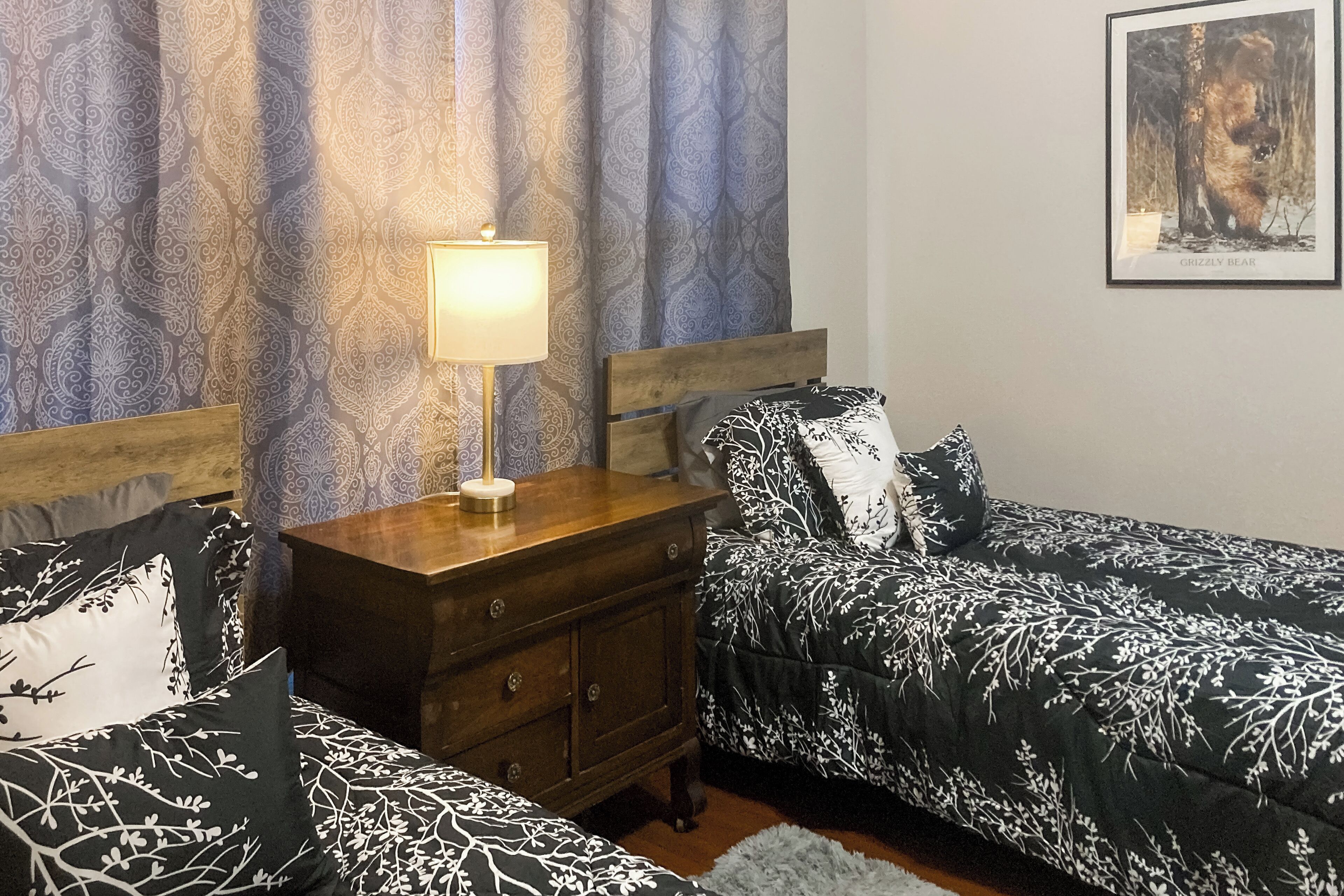 A bedroom with two beds, a nightstand, a lamp, patterned curtains, and a framed picture on the wall.