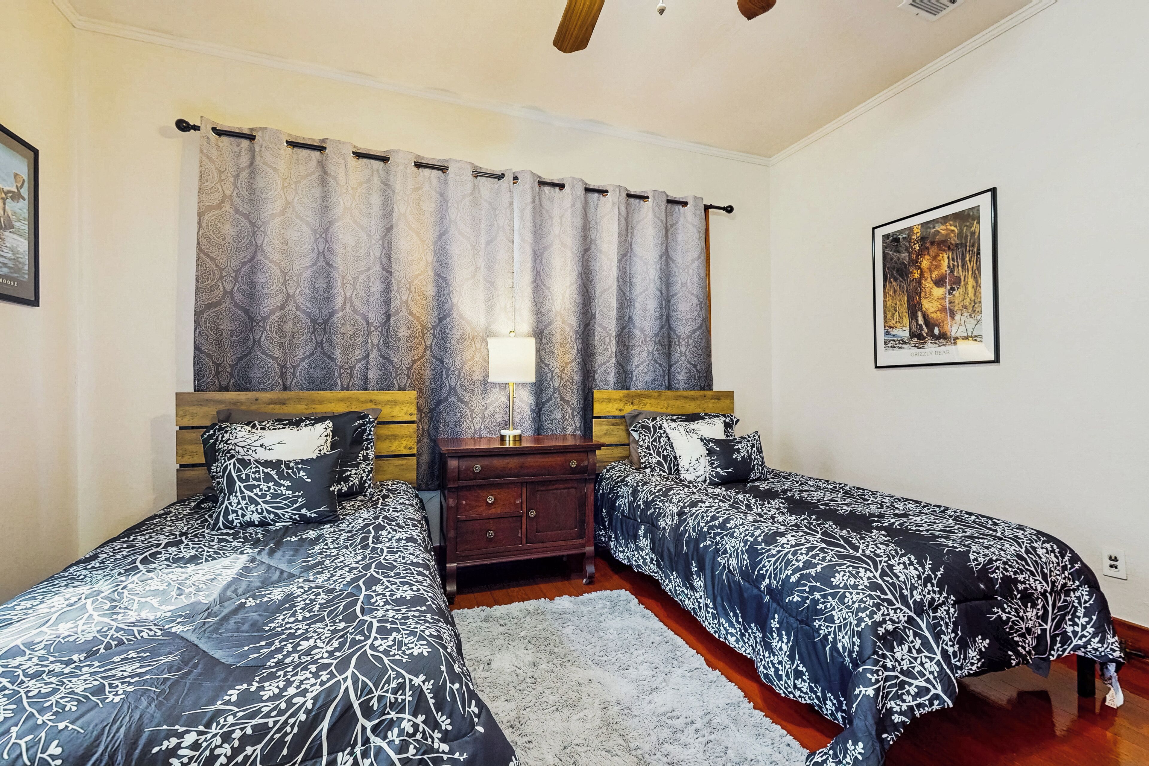 A bedroom with two single beds, a wooden headboard, a nightstand, a lamp, a framed picture on the wall, and a patterned curtain.