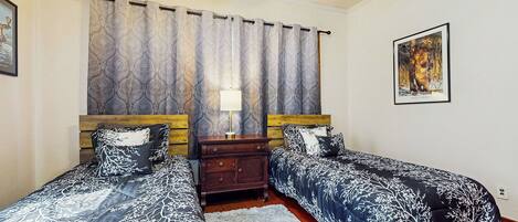 A bedroom with two single beds, a wooden headboard, a nightstand, a lamp, a framed picture on the wall, and a patterned curtain.