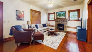 A living room with a sofa, armchair, coffee table, fireplace, and artwork on the walls. - Walkable 1900s Home w/ Porch in Downtown Buda (Buda)